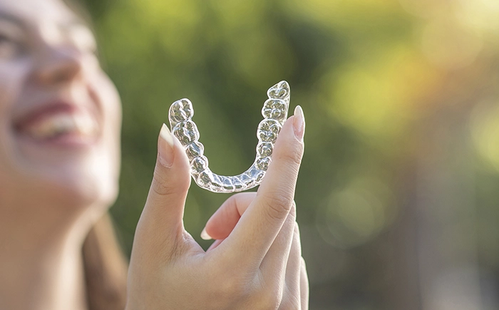 Woman smiling in background while holding a clear retainer in the foreground