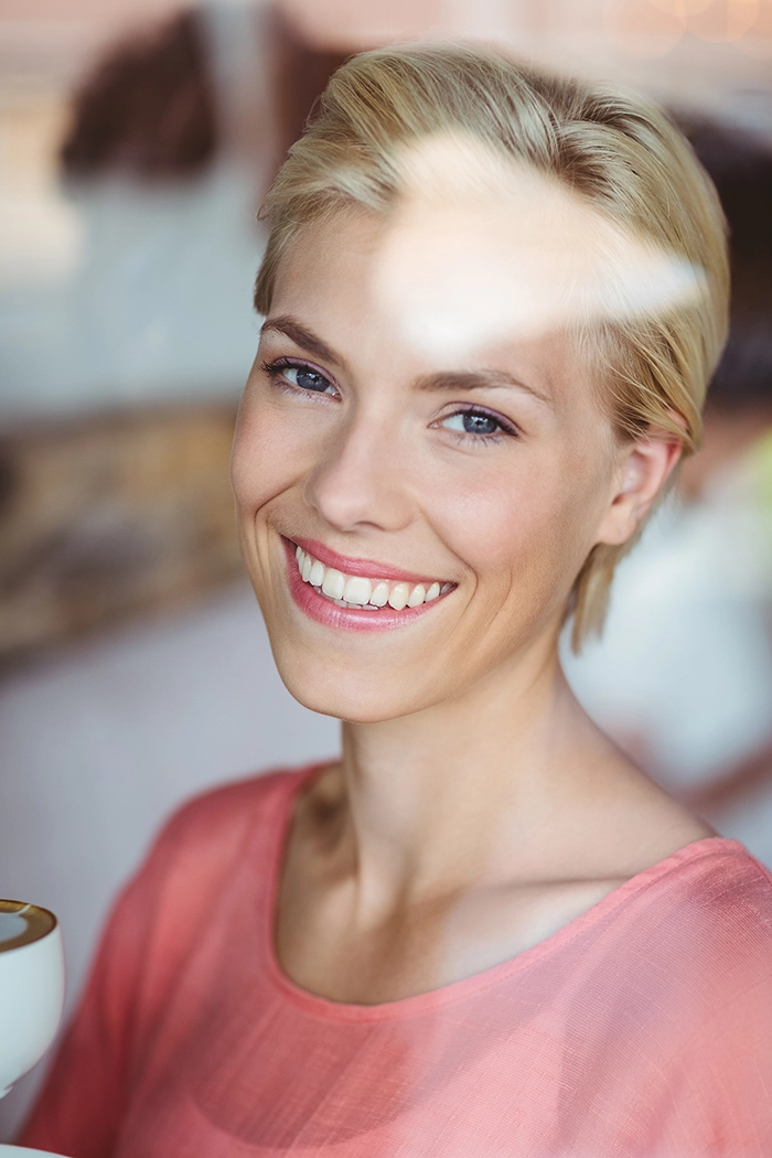 Woman with short blonde hair, wearing a pink shirt and smiling behind a window.
